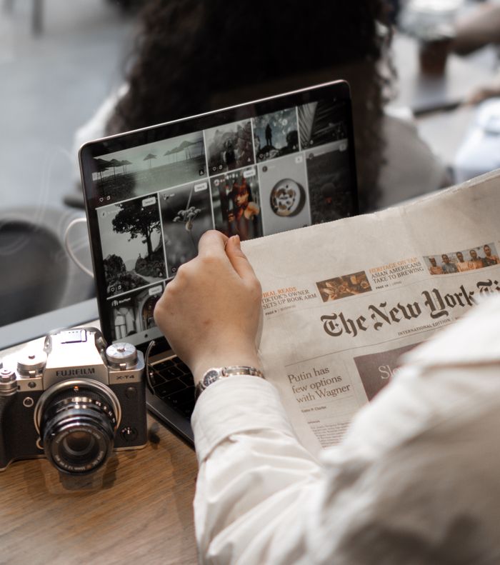 services-04 A person multitasks by reading a newspaper and using a laptop, surrounded by a vintage camera in a cafe setting.