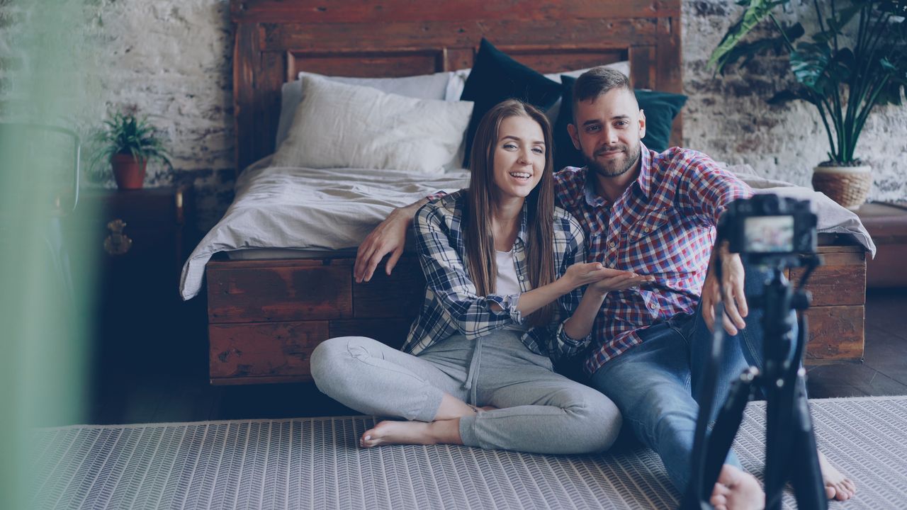 A young couple sits together in a cozy bedroom, filming a vlog with a camera on a tripod.