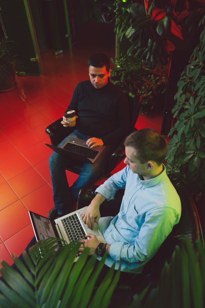 Two men collaborating on laptops in an indoor garden with coffee.