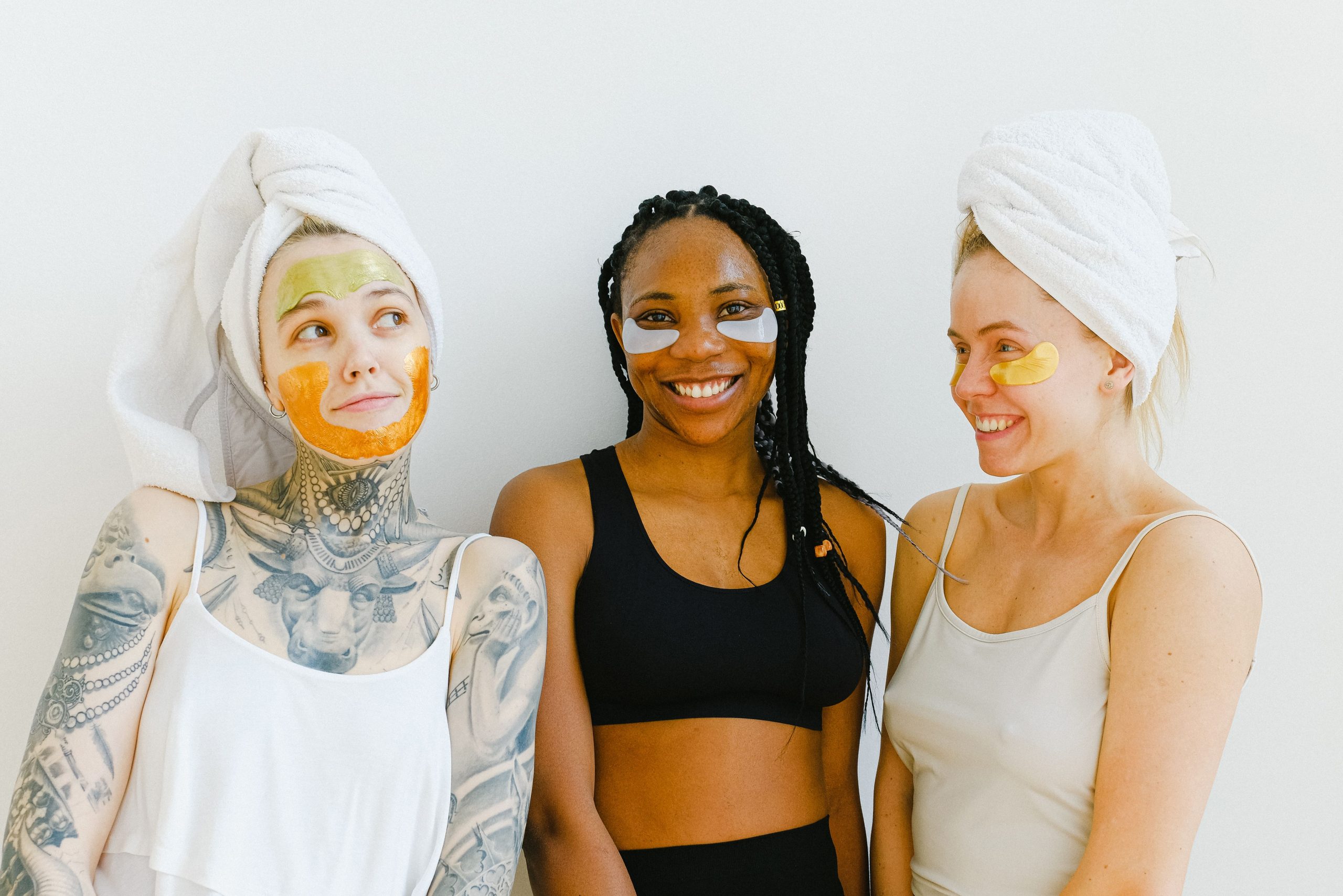 Positive women smiling while having skincare procedure with eyes patches and masks during spa in studio at weekend together near white background